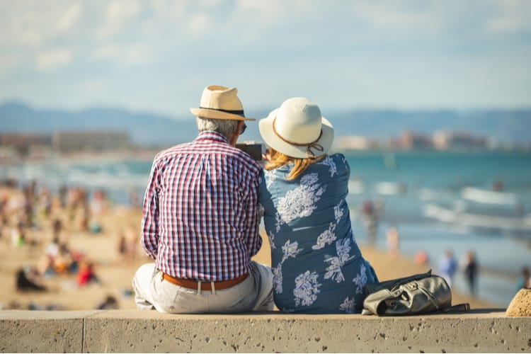 An older couple sitting on a wall at a beach.